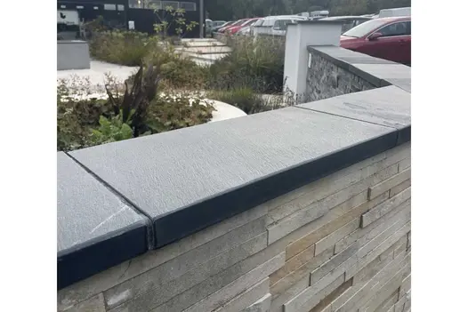 Angled stone-clad wall topped with Brazilian Black slate coping stones divides parked cars from paved area with planting.