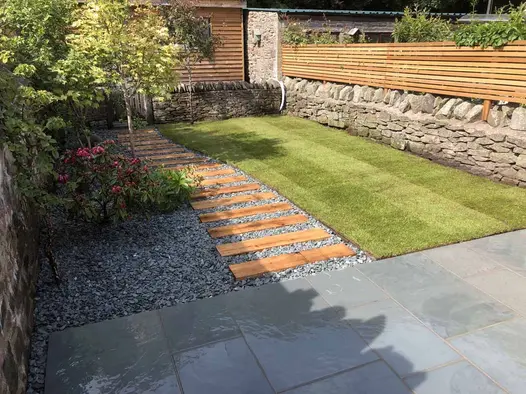 Wood planks set in gravel lead from Brazilian Grey slate patio, past lawn to gate in dry-stone wall, giving entry to garden office.