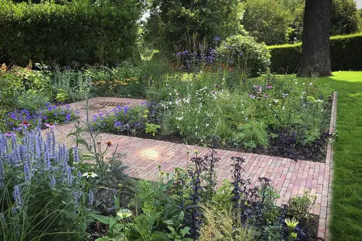 Rectangular area cultivated with flower beds around Seville clay paving path, by hedge in large lawn with mature trees.