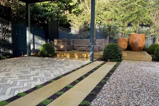Gromo Antica grey brick paving under pergola, in paved garden surrounded by trees, with raised patio. Built by Aoba Landscapes.
