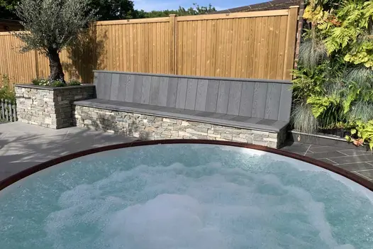 Large round bubbling hot tub sits on patio in front of long bench with seat and back of Brushed Basalt Millboard decking.