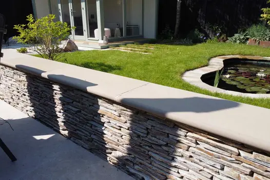Close up of rock wall with bullnose buff sawn sandstone atop also shows lawn and pond in the background.