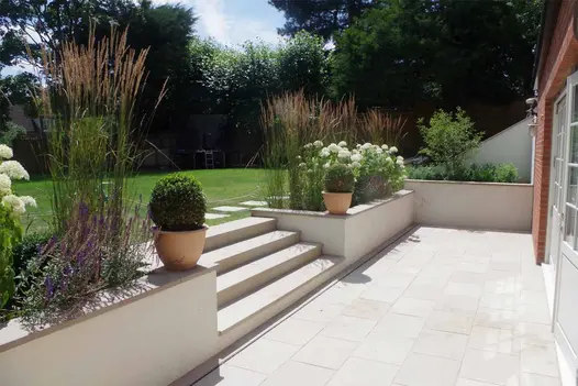 Cream walls edge patio of Buff sawn sandstone, with 4 steps up to stepping stone path in lawn surrounded by bushes.