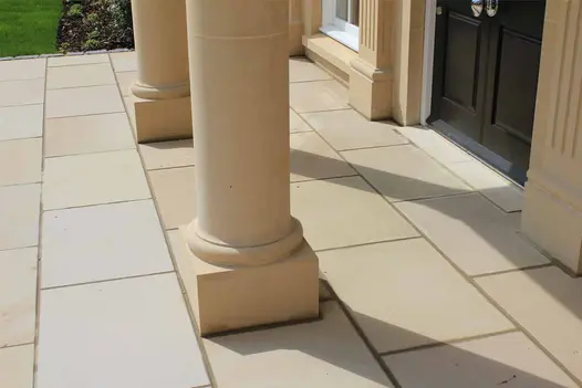 2 square-based stone pillars stand opposite Corinthian doorposts on Buff sawn sandstone paving laid with running bond pattern.