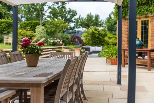 Wooden dining furniture sits under pergola on patio of Buff Smooth sandstone paving. Steps up to lawn and seating area beyond.