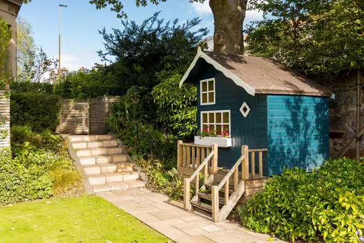 Huge Wendy house in hedged border has steps down to path of Buff Smooth sandstone paving leading to 7 steep steps up to fence.