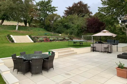 Huge multi-levelled garden shows beige sawn sandstone bullnose steps lead from patio to lawn, and then up a hill to a trampoline.
