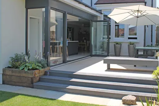 3 wide steps of Millboard bullnose boards rise to patio with matching bench, tall planters and parasol outside bifold doors.
