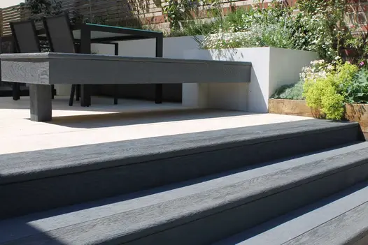 Steps of Millboard decking boards with bullnose edge rise to patio with matching bench attached to raised bed at one end.
