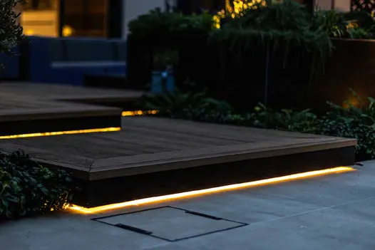 LED strip lights luminate burnt cedar millboard decking with other lights in the background between foliage.