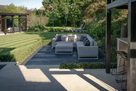Sun casts shadow over burnt cedar millboard decking area, hosted by large corner outdoor sofa with well manicured lawn in the background.