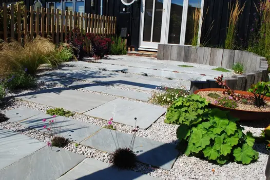 Sunny back garden paved with Kandla Grey sandstone slabs of different sizes, set into gravel with dry planting. 