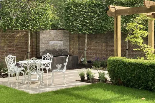 White dining set on Faro porcelain paving edged with water feature, pleached trees and pergola. Built by Bushy Business Gardens.