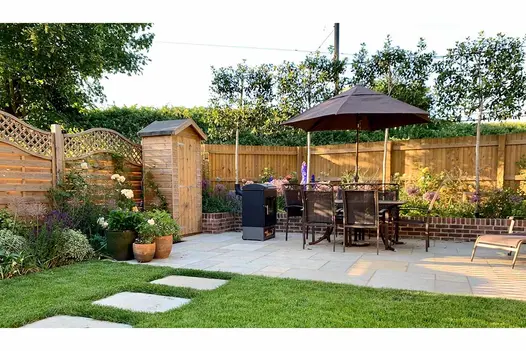 Kota Brown limestone slabs set into lawn lead to matching oblong patio with dining set, tall tool shed and raised bed.