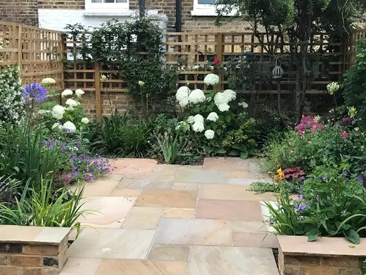 End of narrow urban garden with wall and trellis on 3 sides of Camel Dust Indian sandstone paving edged with planted borders.