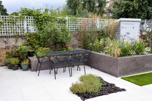 Dining table sits on light patio next to overflowing raised flowerbeds clad in vulcano ceniza porcelain cladding.