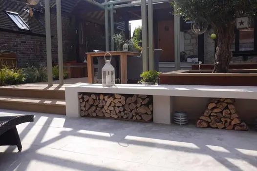 2 steps rise from Jura Grey smooth paving to seating area with grey pergola, table and olive tree in raised planting pit.