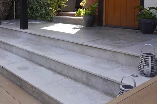 3 wide steps rise from decking to narrow, shadowy  paved area, all laid with Jura Grey smooth limestone paving slabs.