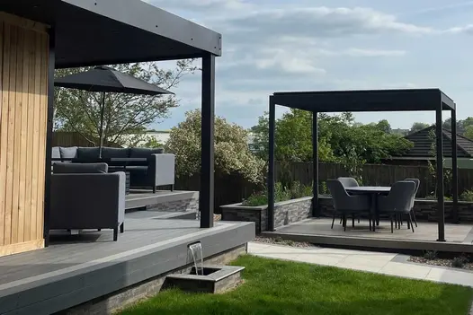 Proteus Grey aluminium pergola on paved area bounded on 3 sides by low wall and path. Garden building with verandah in foreground.