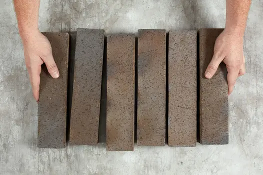 2 hands hold a paver at either end of row of 6 Chelsea Belgium Bricks laid on-edge, showing colour variation and texture.