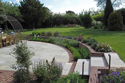Lawn edged with curved retaining brick wall. 3 steps descend to Kota Brown limestone patio with clay paver detailing.