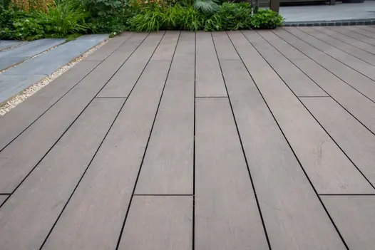 Garden room with planted pots has doors open onto Chestnut Brushed DesignBoard Garden decking edged by foliage and greenhouse.
