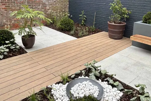 Composite decking runs through patio of light grey porcelain paving with flowerbeds surrounding.