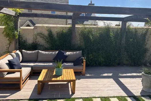 Sun shines thorugh wooden pergola that hosts corner outdoor couch and table sitting on cinnamon designboard composite decking.