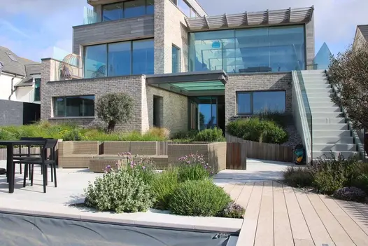 Rectilinear modern house with paved exterior and outdoor furniture. Steps up to 1st floor from decking in Cinnamon DesignBoard.