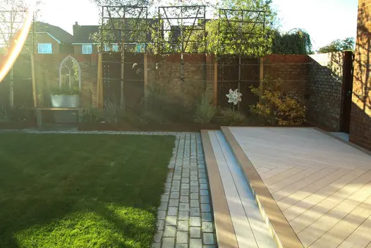 Granite setts edge lawn at base of 2 wide, shallow steps up to decking of Cinnamon composite planks laid in herringbone pattern.