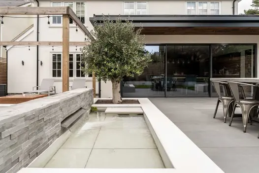 Wide spout in low wall of Kandla Grey stone cladding pours water into oblong pond with porcelain tiles. Design by Gadsden Gardens.