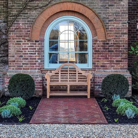 Symmetrical design of Lutyens bench beneath arched window of house with apron of Old English clay pavers between 2 formal beds.