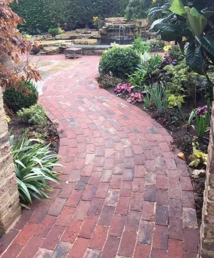Sinuous path of Old English clay pavers runs between planted beds towards rocky water feature in front of hedge. 