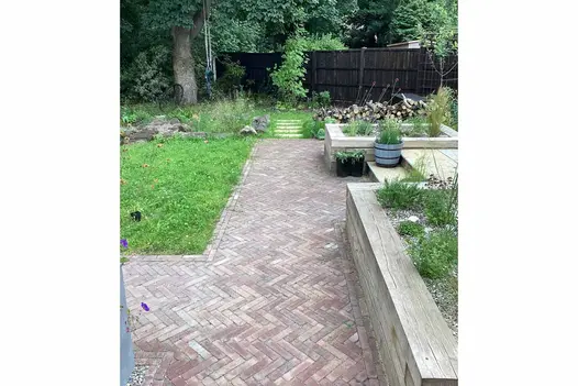 Stone steps descend from patio between 2 timber raised beds to wide Bexhill clay paver path which edges small lawn.