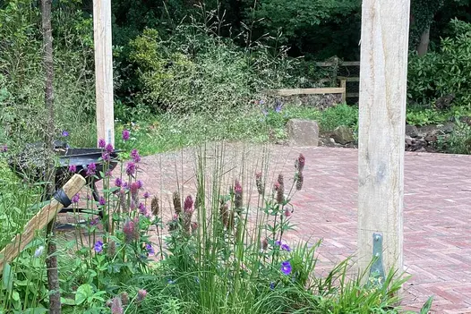 Metal barbecue part hidden behind airy planting by large empty area of red and grey Bexhill clay paving, with stile on far side.
