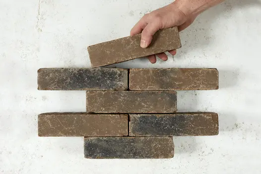 A hand laying a 7th Aldridge clay paver to 6 existing ones on a white background, illustrating the easy placement and natural design.