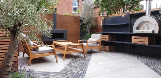 Porcelain paving with diagonal strips of Silver Grey Multi brick paving by shelves and pizza oven against slatted fence.