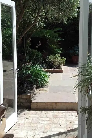 View through open french doors to brick pavers with step up to Tumbled Raj Green sandstone paving and beds with shrubs and trees.