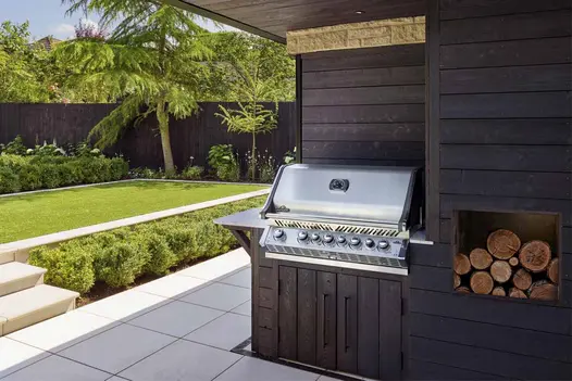 Outdoor cooking area with bbq and wooden log storage overlooking a Comblanchien porcelain patio with steps by London Stone.