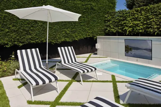 Outdoor TV wall above swimming pool, sun-loungers and parasol nearby, with surrounding area paved with Comblanchien patio tiles.