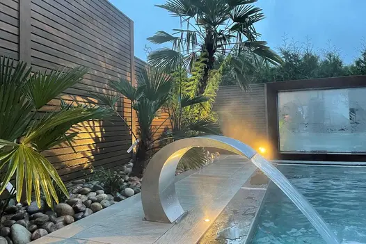 C-shaped water feature shoots flume into swimming pool in front of pebble-mulched bed at base of fence of Warm Teak composite wood.