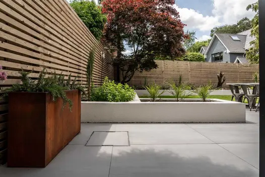 Corten steel tall trough planter filled with spiky plants stands on paving in front of open slatted wooden fence.