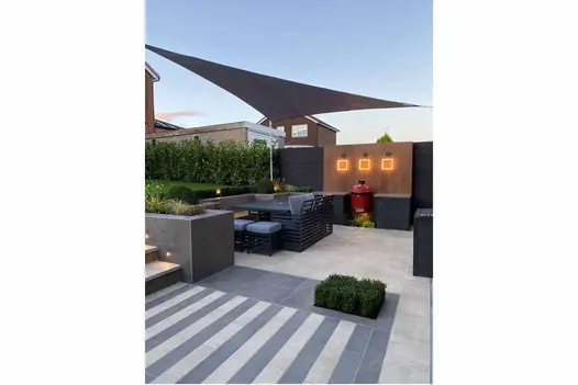 Alternate steel-grey and cream porcelain planks lead up to steps from patio, with outdoor kitchen and dining set, to lawn level.