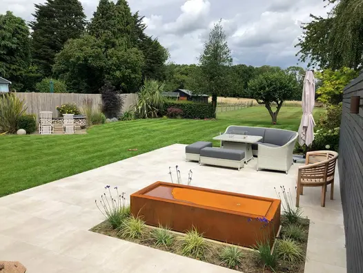 Garden furniture and corten steel water feature sitting amidst a cream porcelain patio at the top of large garden backing onto meadows.