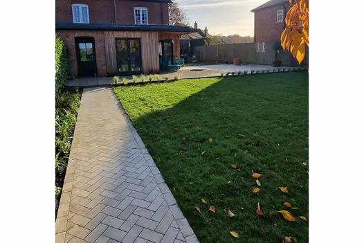 Path of Cream porcelain setts leads down side of lawn to back of house with wood-clad extension and patio wrapped around side.