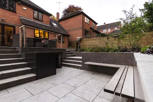 Two sets of steps descend from suburban house to silver-grey porcelain patio, enclosed by raised beds. Design by Creative Roots.