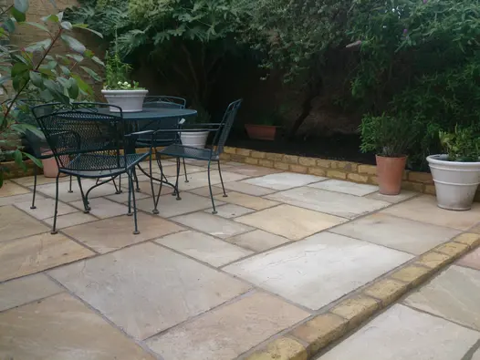 Angled shot of a garden corner featuring black metal furniture on tumbled mint sandstone paving, with a tall bush on the right.
