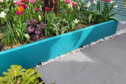 Distinctively coloured turquoise raised bed planted with flowers and annuals surrounded by Dark Grey porcelain paving.