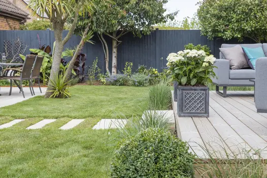 Wooden deck with step down to path of Beige smooth sandstone plank paving slabs set into grass, leading to paved area with table.