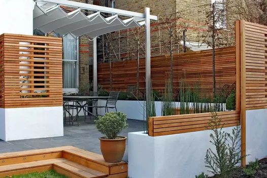 2 wooden steps descend from Midnight Black limestone patio with pergola and edged with slatted fencing. Design by David Simpson.